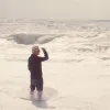 Ludovico Einaudi stands knee-deep in the sea facing away from the camera, holding his hat with one hand.