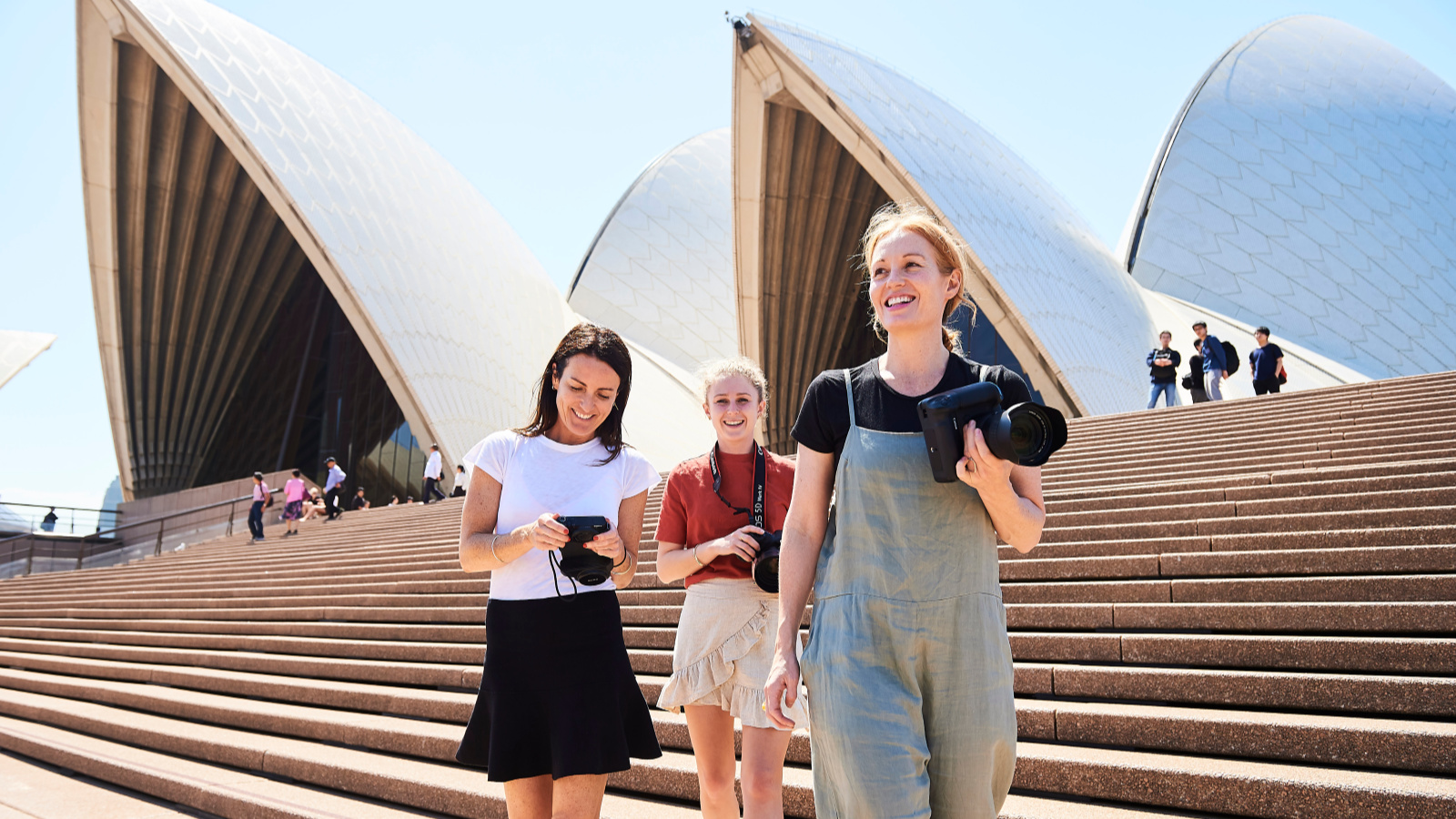 Three women with cameras walking down the steps of the Sydney Opera House.