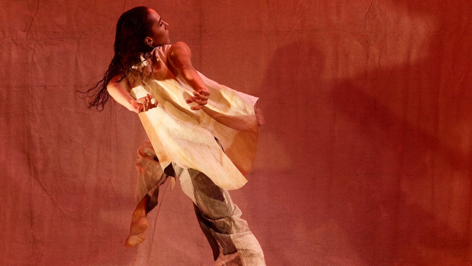 The back of a dancer in mid movement with one leg lifted and hands thrown back. Head looking to one side, against an ochre coloured backdrop.