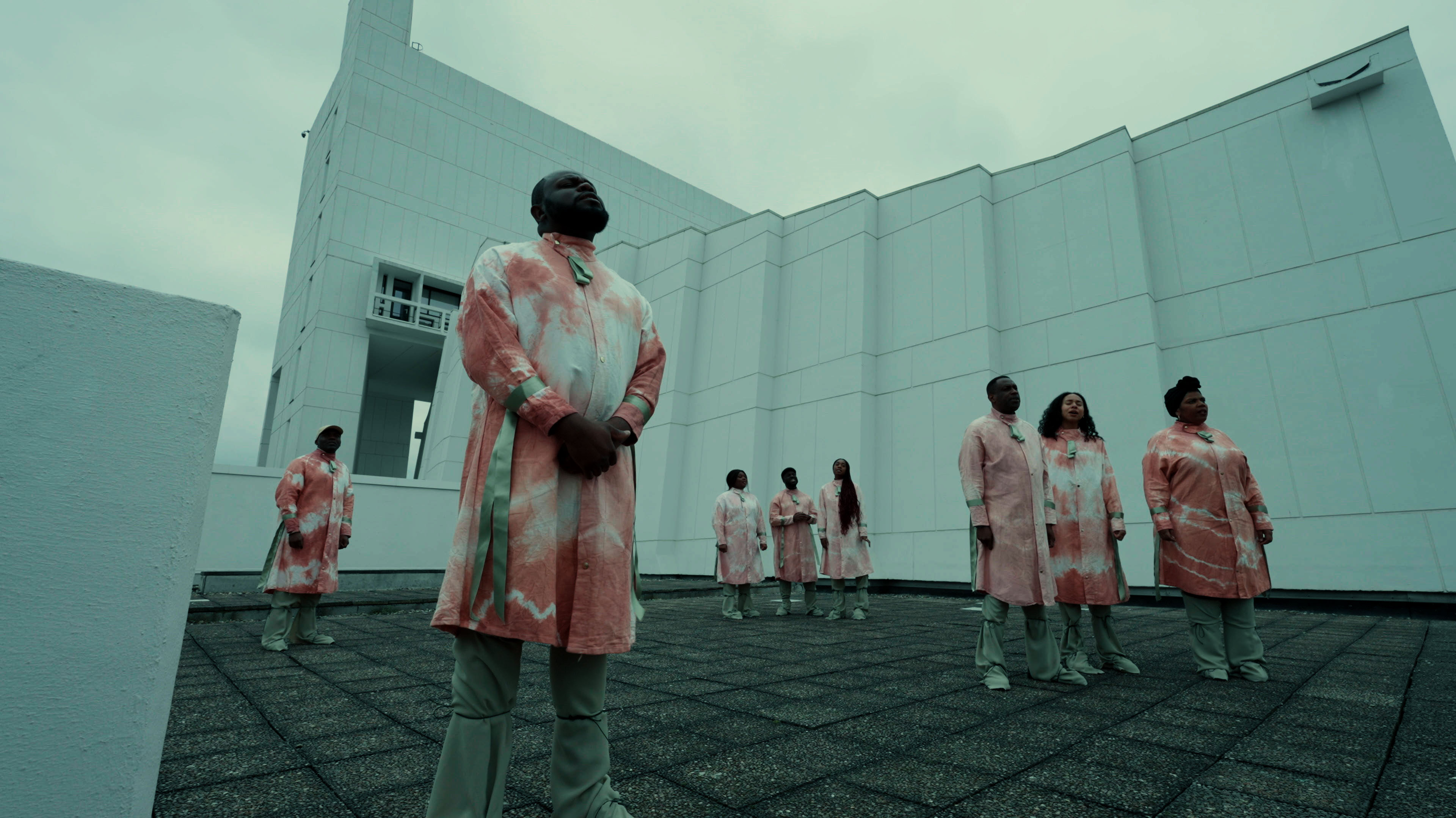 A group of black people are standing spread out on a balcony on a cloudy day, wearing red and white tie dye clothing.