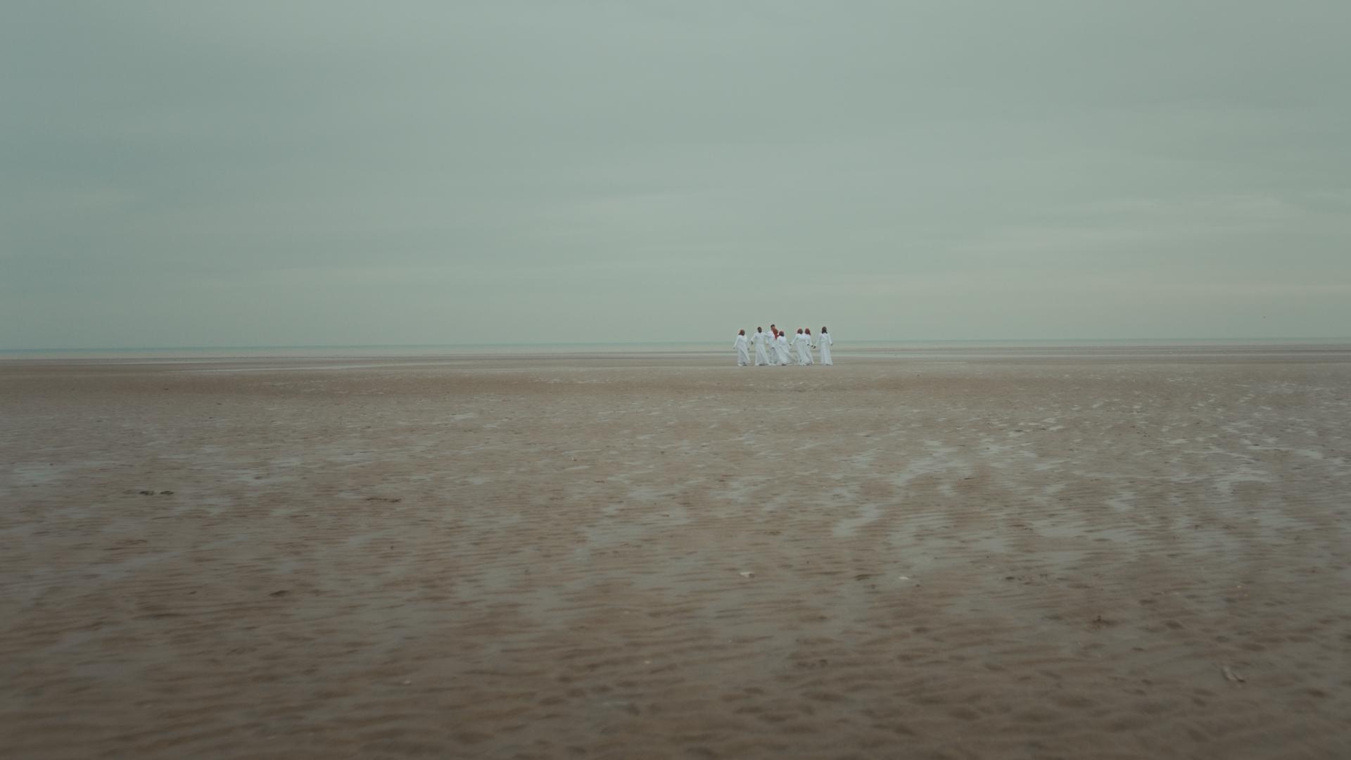 In a barren desert with a grey cloudy sky, a group of people stand in the distance in all white clothing.