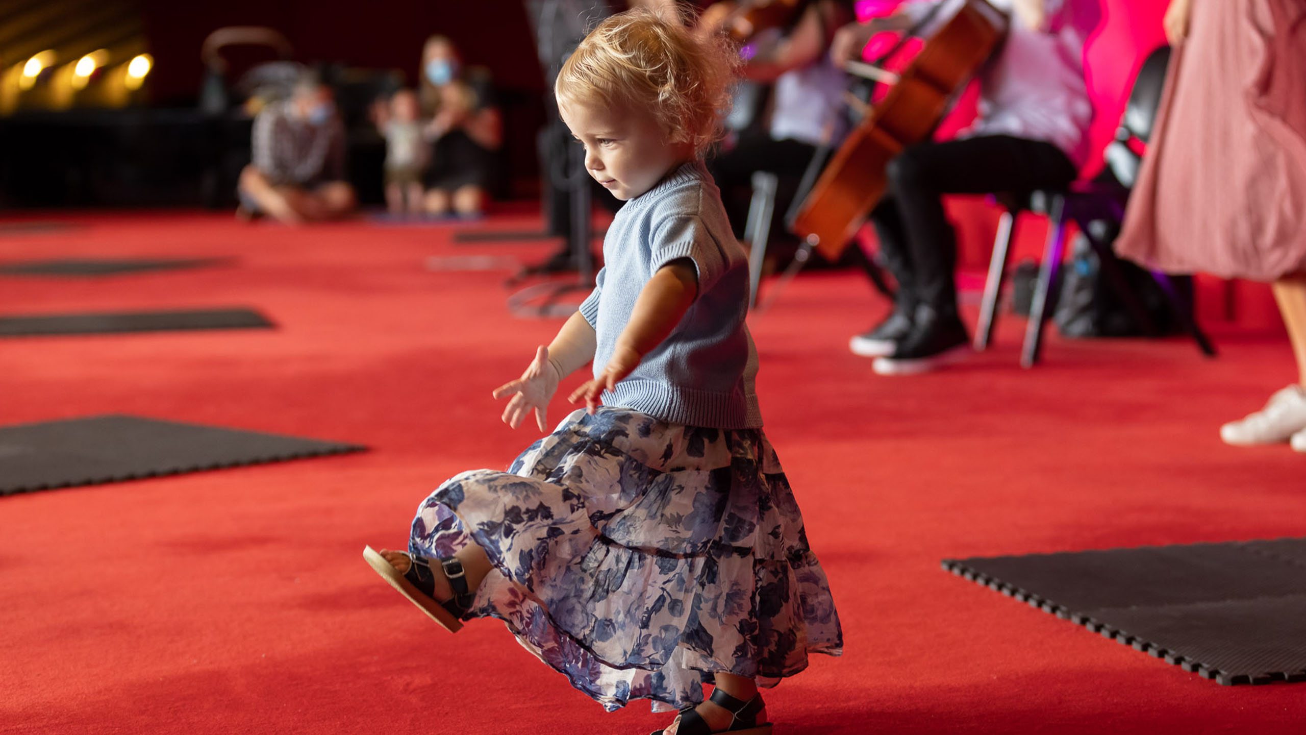 A toddler in a blue sweater and floral skirt dances on a red carpeted floor with people and musicians playing string instruments in the background.