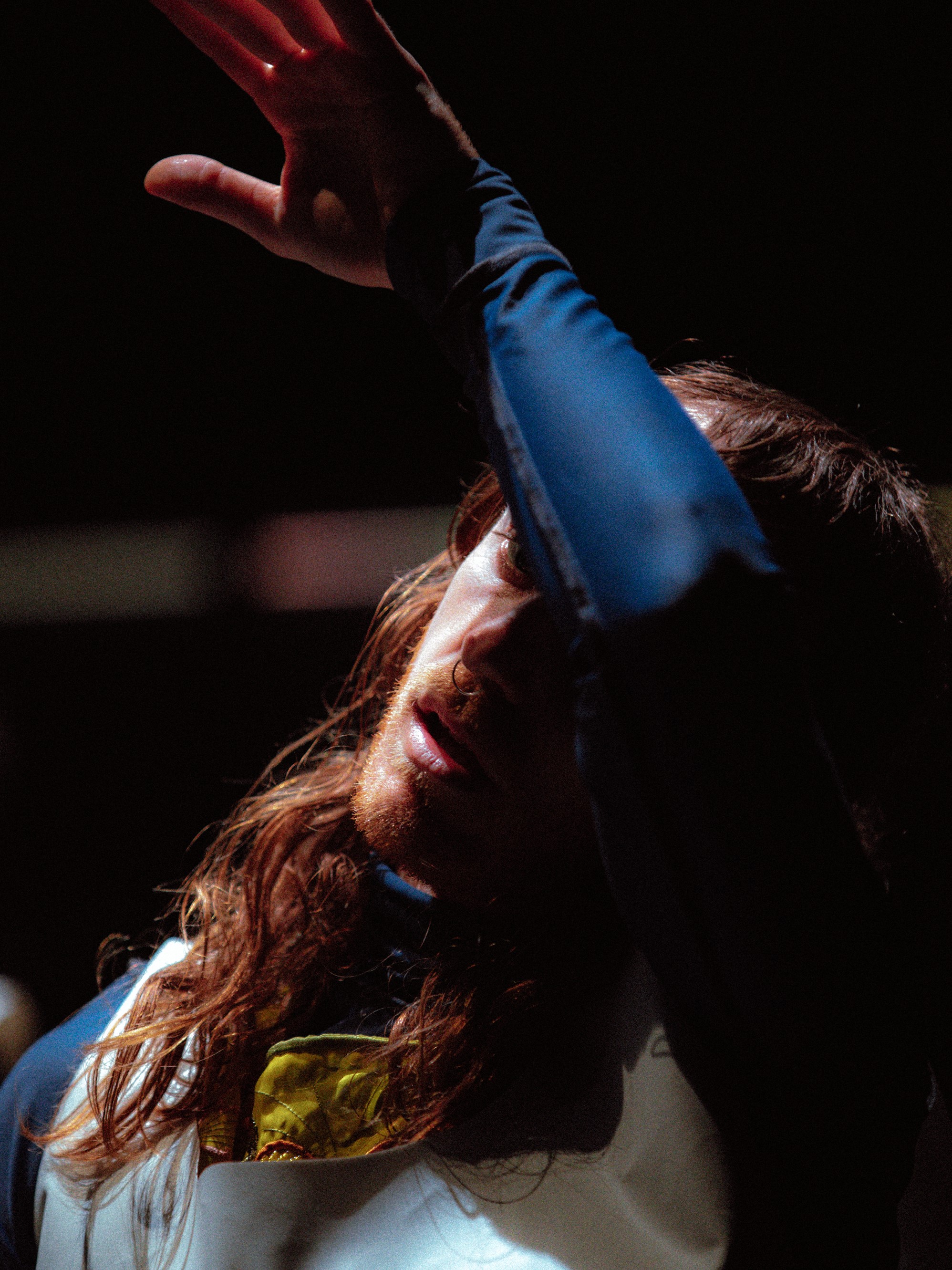 A man with long hair poses thoughtfully mid-movement, he wipes his forehead with his forearm.