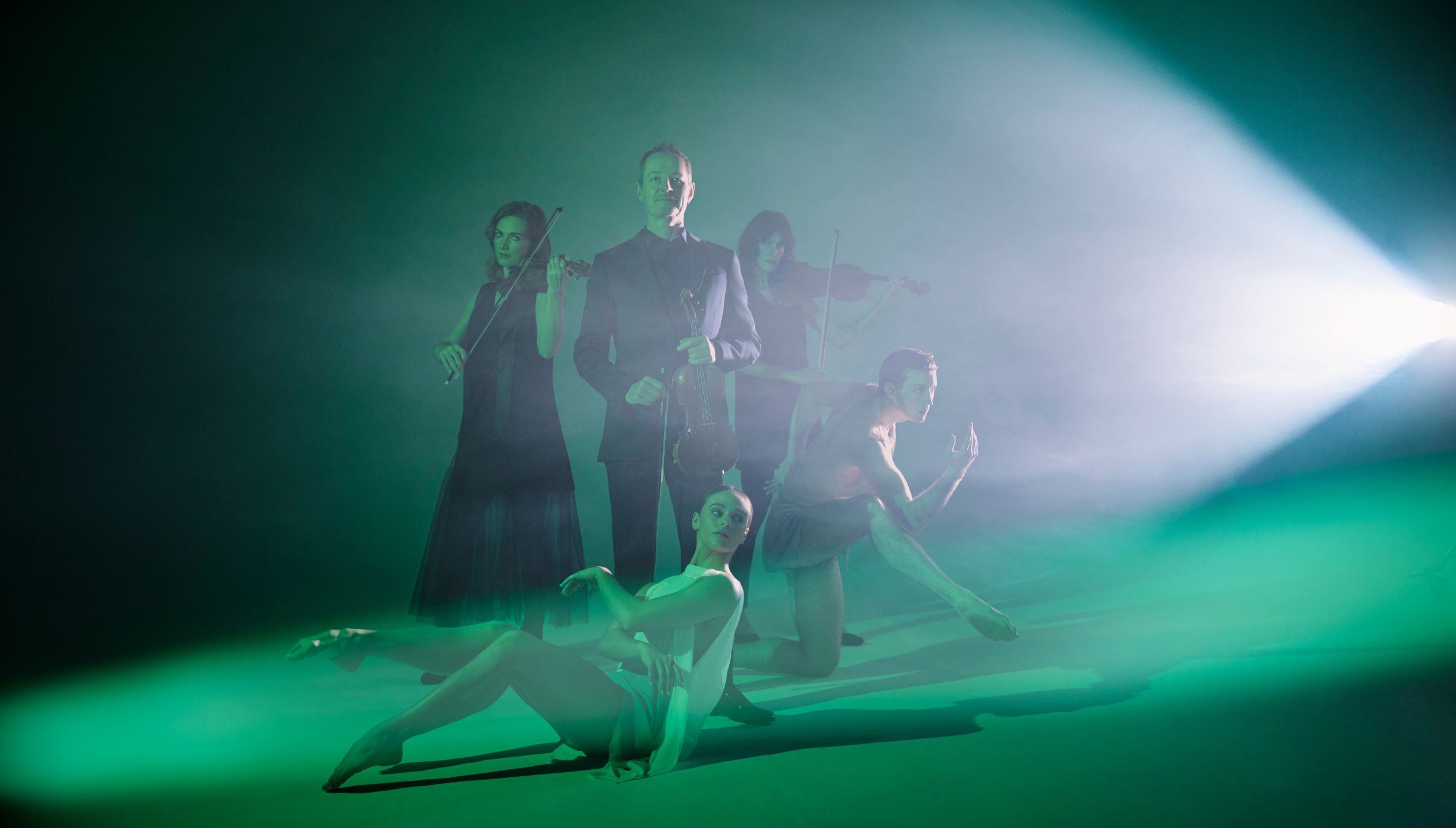 A white, 50-year old man holds a violin immersed in green light. Around him is two female violinists, a man dancer and a female ballerina.