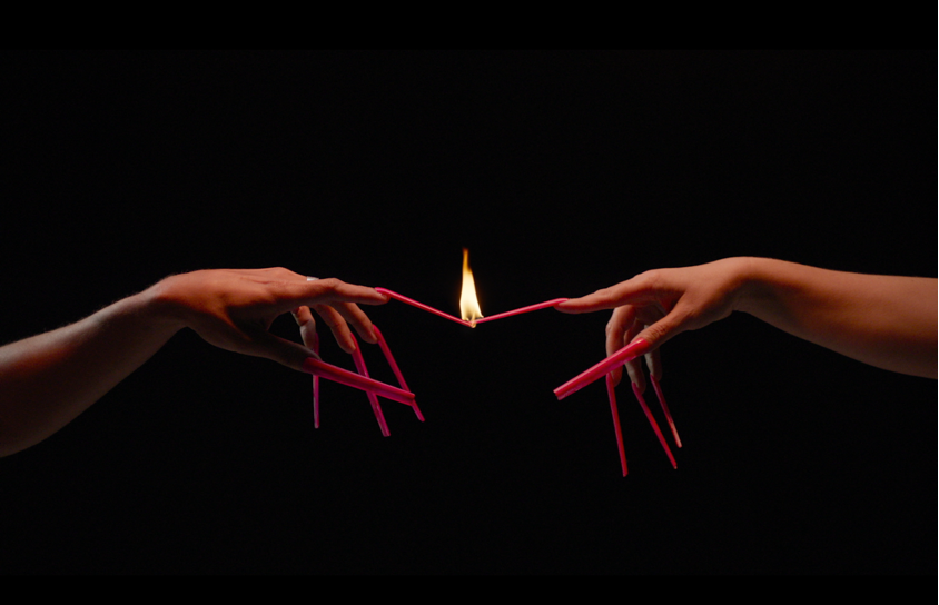 Two hands with long pink acrylic nails point at each other with their index fingers. A small yellow flame sits where the fingers meet.