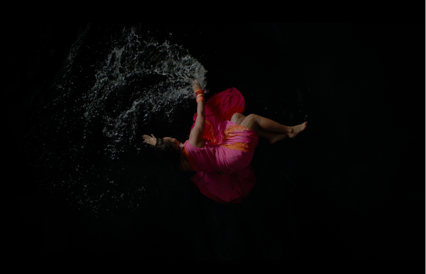 A birdseye view of a white woman in a pink and orange dress laying in shallow water on a black ground.