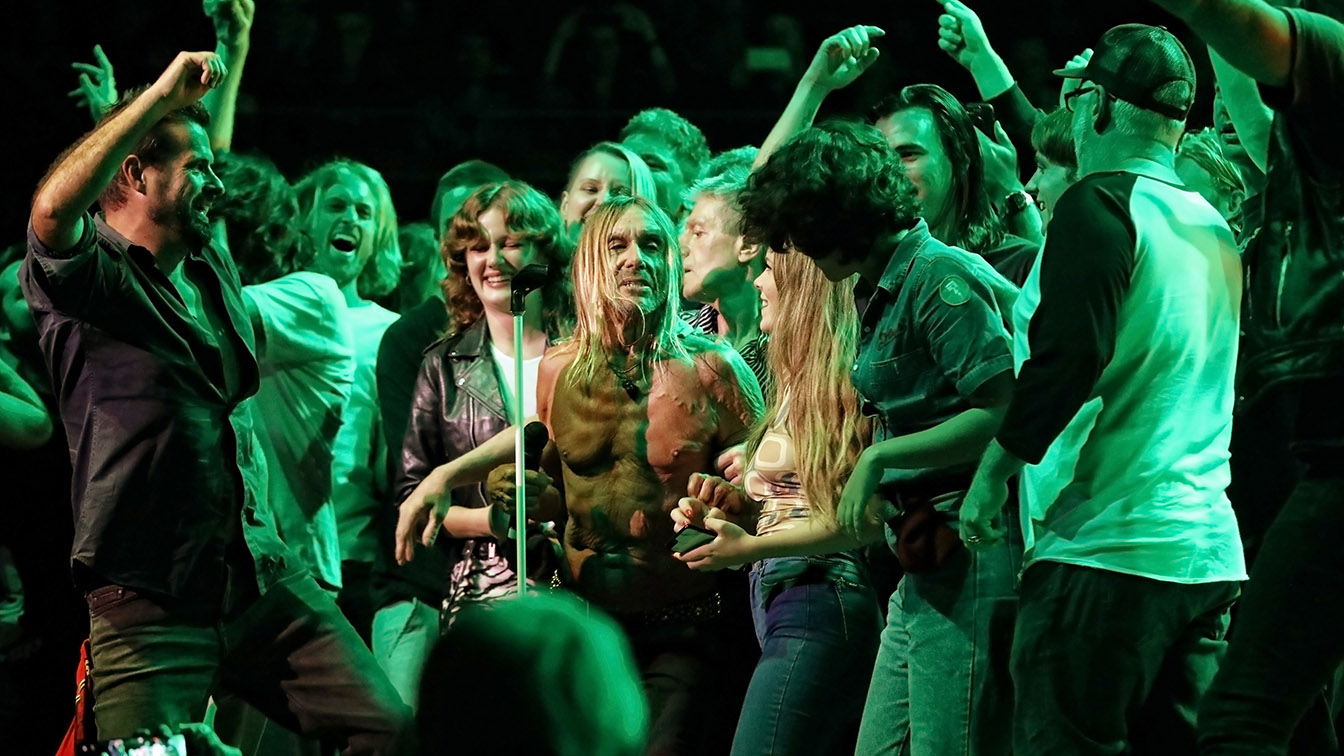 A topless man with long blonde hair singing into a microphone, surrounded by a green-lit lively crowd.