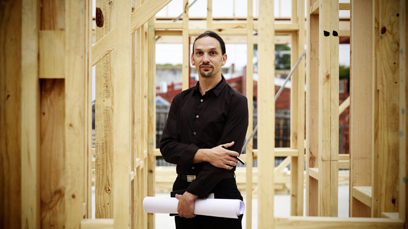 A man in a black shirt and long hair standing underneath wooden construction work.
