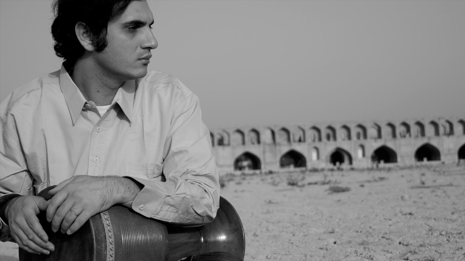 A man in a shirt sat down on sand, holding an instrument.