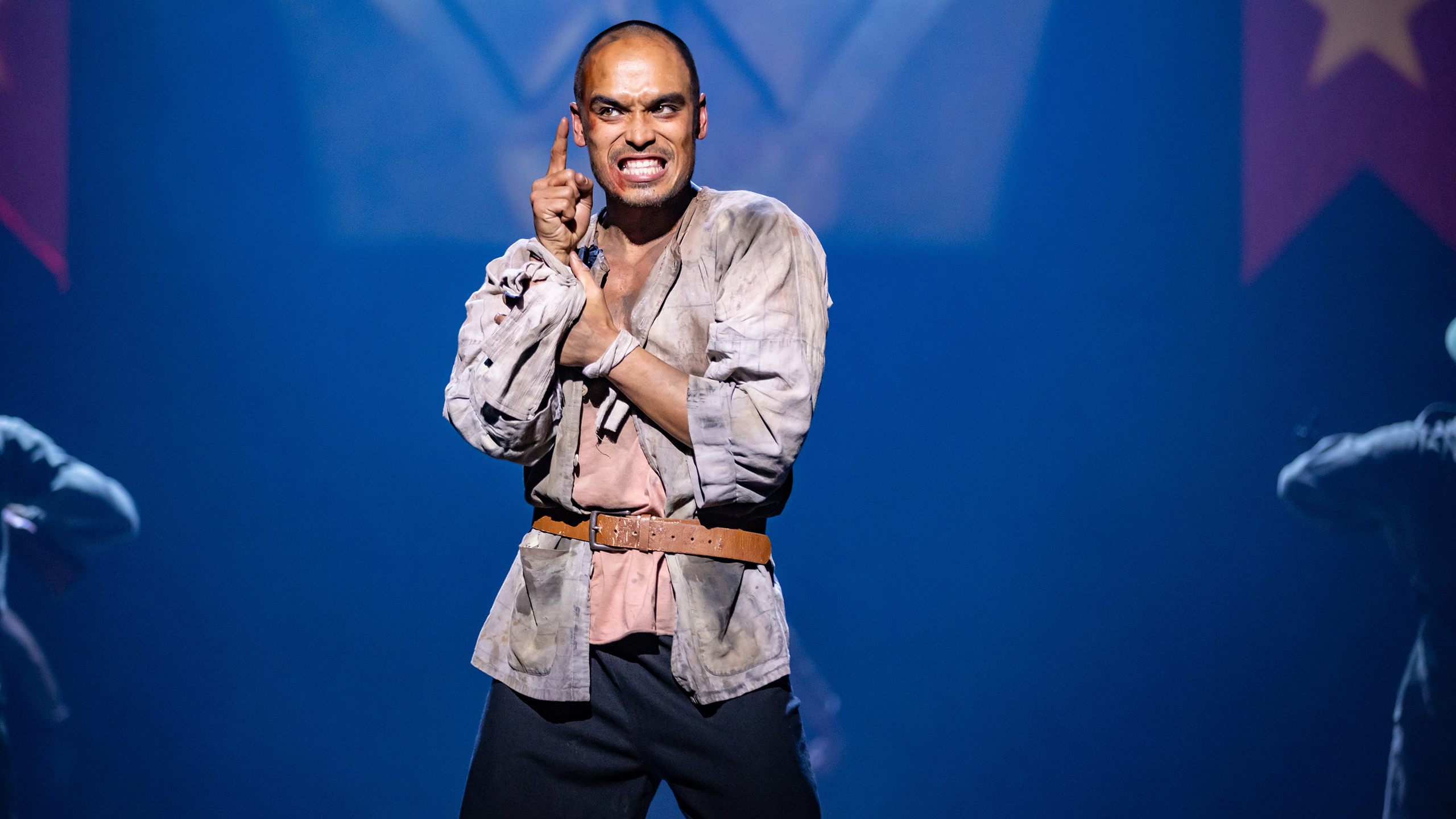 An Asian man with tanned skin and shaved hair wears a shirt with dirt on it and a belt. He is singing.