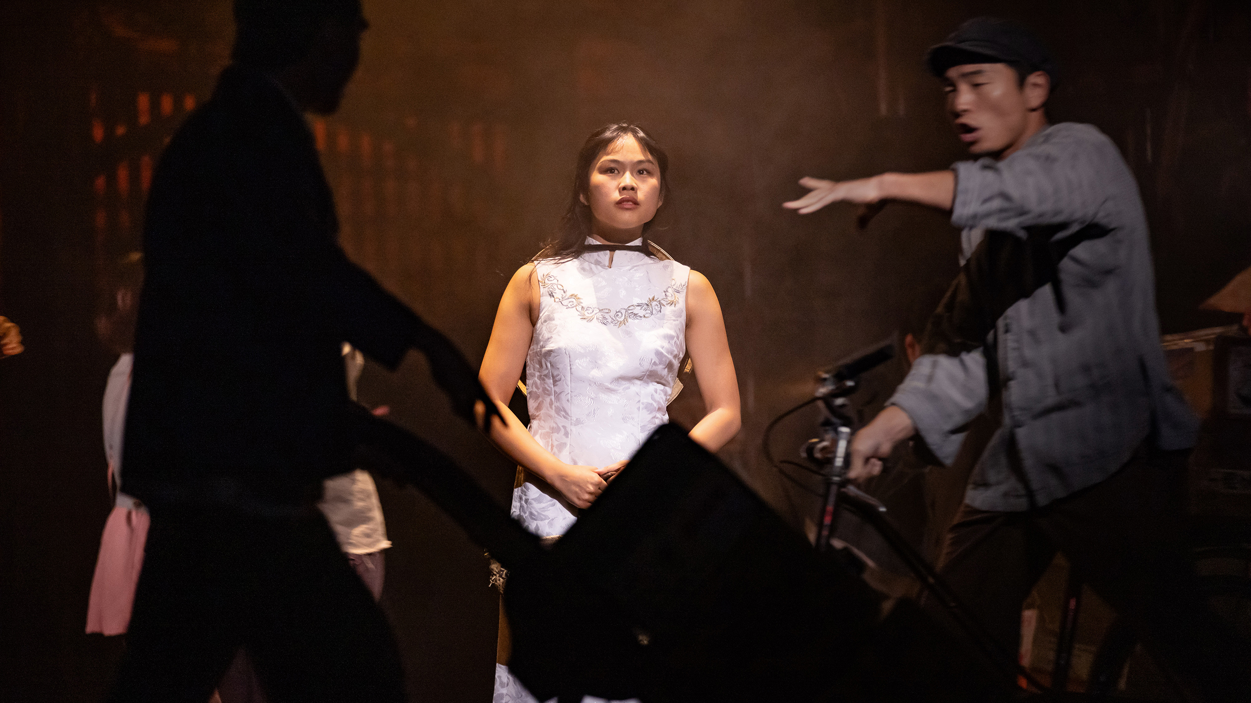 An Asian woman wearing a traditional white dress stands centre stage while cast members move around her.