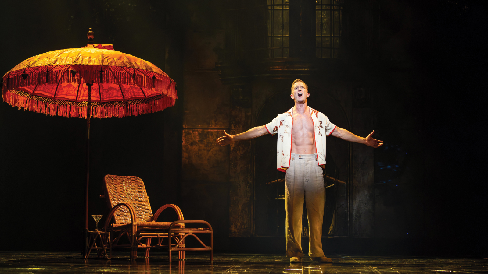 A man stands on a dimly lit stage with arms outstretched, singing. He is shirtless under an open button-down shirt. A wicker chair and a red umbrella are beside him.