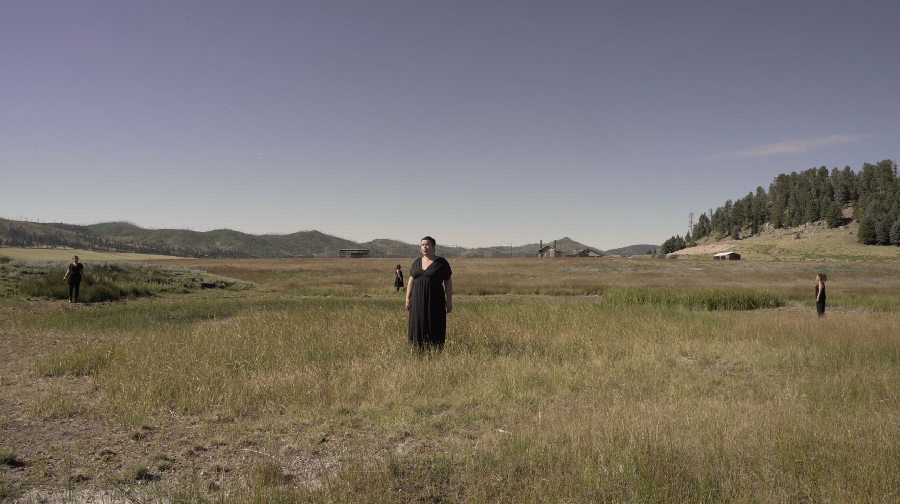 A white woman with short dark hair wearin a black dress stands in a field with blue skys behind her.