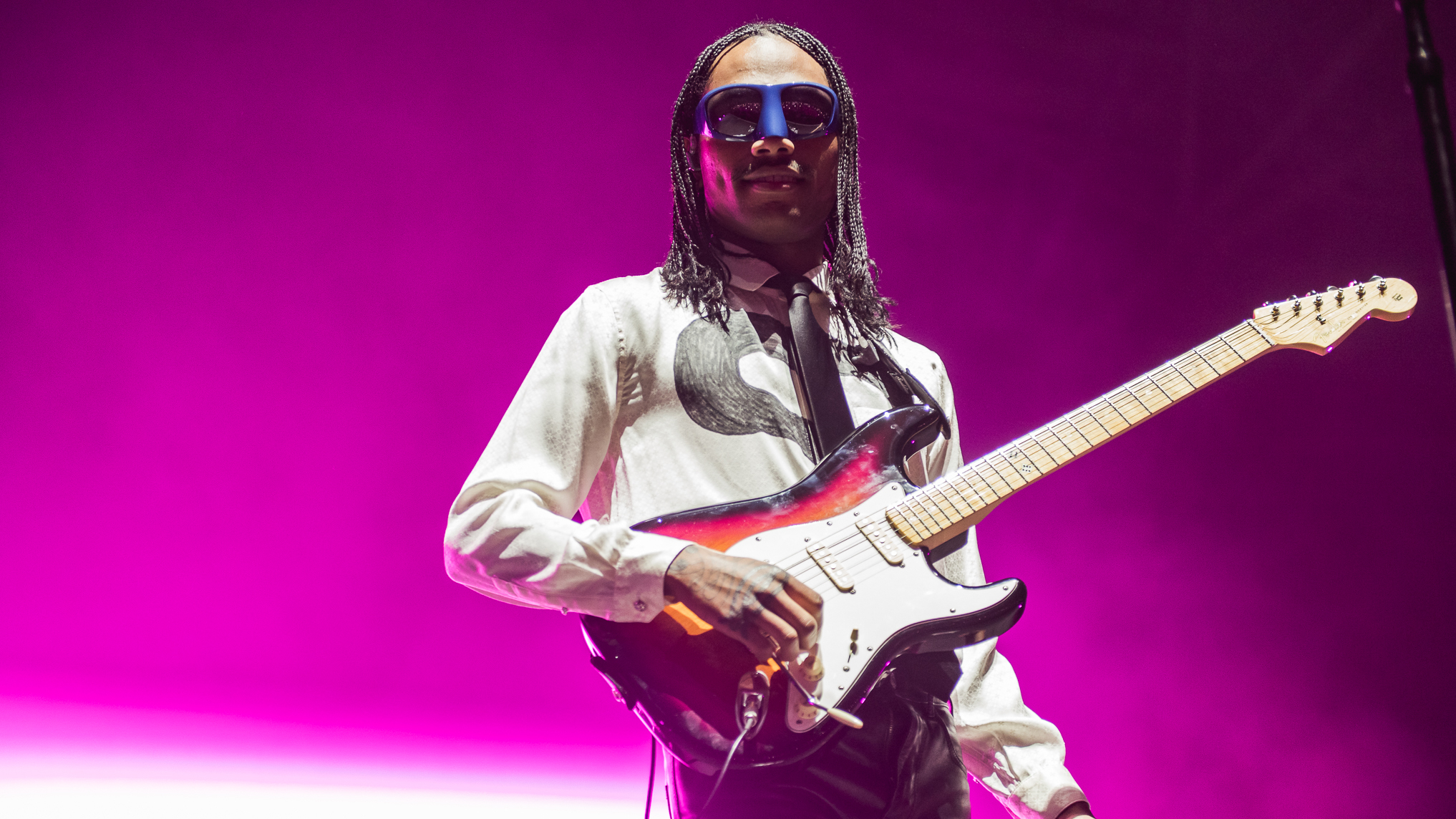 A person wearing googles and a white long sleeve t-shirt playing a guitar against a bright purple background.