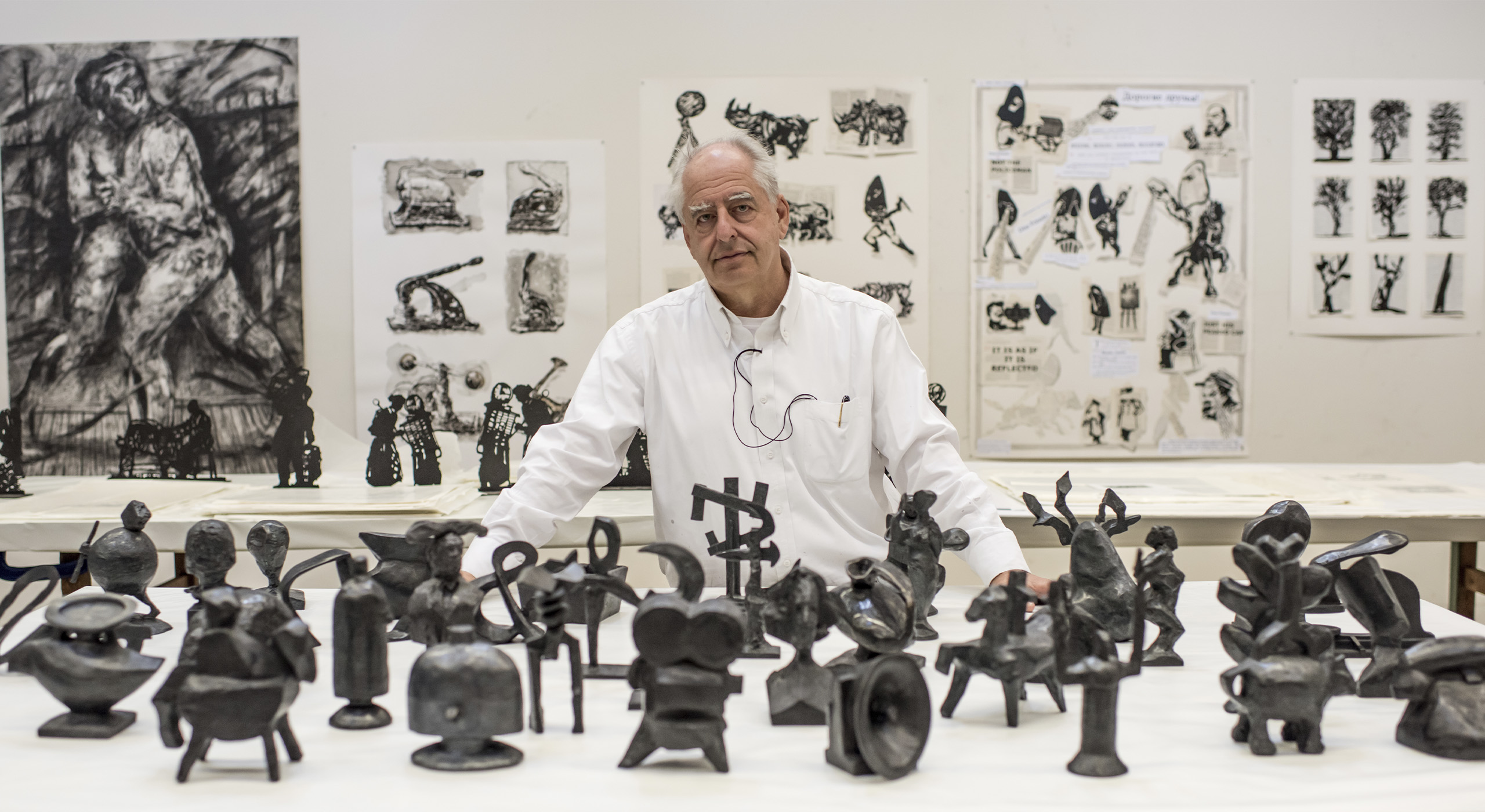 A mature man with white hair stares forward surrounded by details illustrations and sculptures.