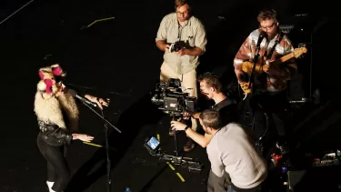 Three men filming a woman singing, whilst a man plays a guitar.