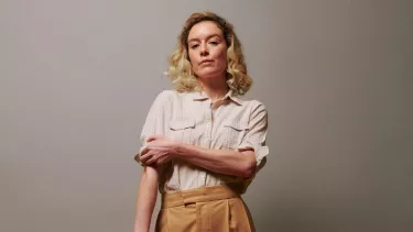 A white woman with blonde shoulder-length hair wears a beige shirt and light brown pants, she's rolling up her sleeves.