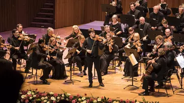 An orchestra performing on stage as viewed from the audience. A violinist is standing centre stage.