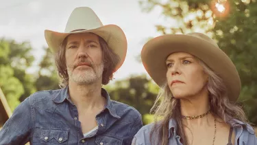 A man holding a guitar and woman both in cowboy hats look pensive away from the camera exuding a country music vibe.