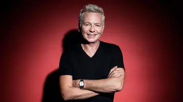 A white man in his 60s with grey hair wears a black t-shirt and has his arms crossed, standing in front of a red background.