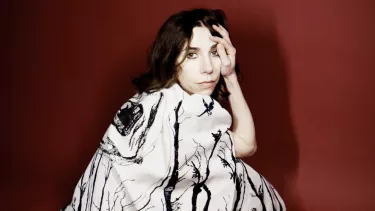 A white woman with long brown hair wears a stylish white dress with a black splash pattern. She's crouched down and looking at the camera.