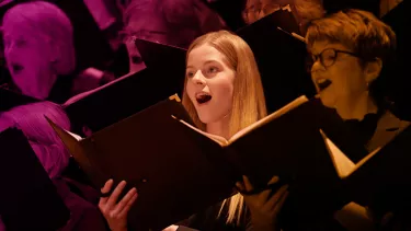 A blonde girl with an open song book sings amongst a choir
