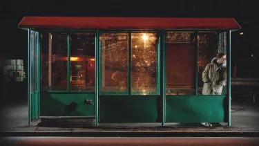 A man in a thick coat leans against an empty bus stop at night.