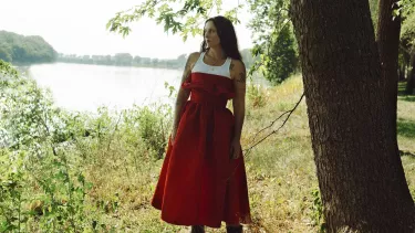 Woman in red dress standing by river, gazing into distance.