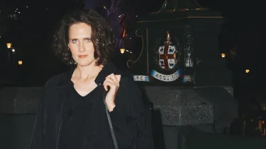 A women standing on a dark street at night, wearing black, with an instrument case over her shoulder.