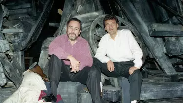 Two men seated on set, with wooden objects behind them.