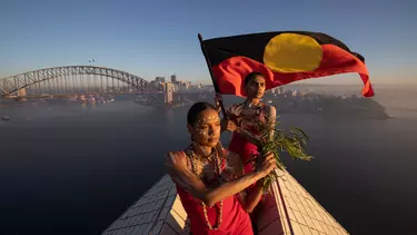 Two women on top of the Sydney opera house hoisting the Aboriginal flag.