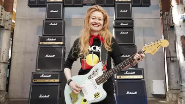 A woman with long hair playing an electric guitar in front of a tower of amps.