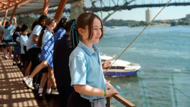 A young girl wearing a school uniform stares out a window in a Sydney Opera House foyer. The Sydney Harbour Bridge is seen in the background.