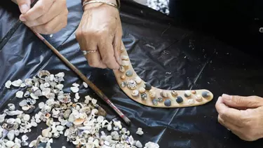 Two hands are placing small shells onto a light brown boomerang.