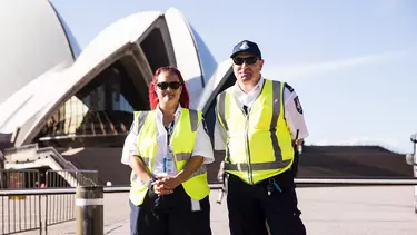 Two security officers wearing high visibility vests stand in front of the Opera House.