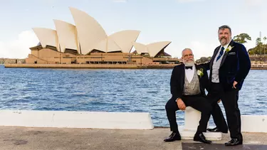Two men stood in suits outside of the Sydney Opera House.