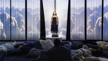 A young white woman with dark brown hair stands in front of a fabric background, where projections of clouds are being cast. An audience of meditation children and adults lie on pillows in front of her.