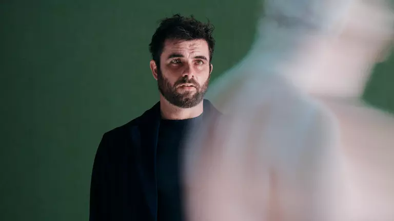 A white man with black hair and dark beard wears all black. He's in front of a green background and is looking at a shadow.