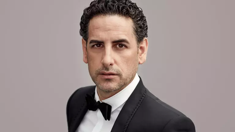 A man with dark curly hair looks at the camera wearing a tuxedo