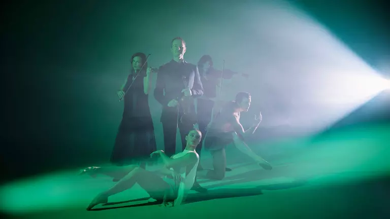 A white, 50-year old man holds a violin immersed in green light. Around him is two female violinists, a man dancer and a female ballerina.