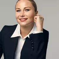 A blonde woman wearing a white shirt with suit leaning on to a chair.