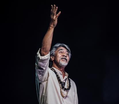 A Pacific Islander man with white hair and a beard stands on a blank stage raising his right arm up.