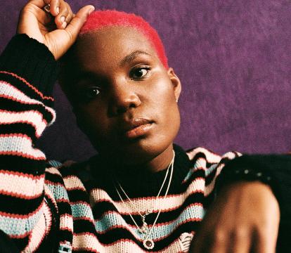Arlo Parks wearing a striped knitted sweater, sitting on the ground against a dark purple wall.