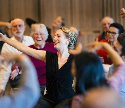 A woman in a black dress smiling, reaching out with an audience surrounding her.