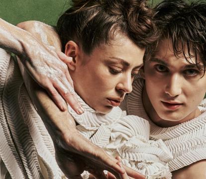 Two dancers in off-white textured costume with arms interlocked and white clay on their bare arms, against a dark green background.