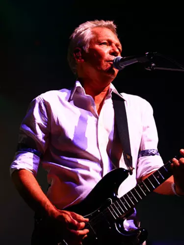 A white man with grey hair is playing an electric guitar and singing into a microphone.
