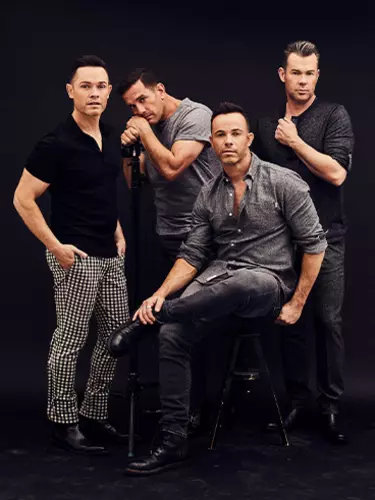 Four white men wearing various shades of black, white and grey are posed in front of a black background. Three men are standing and one is seated on a stool.