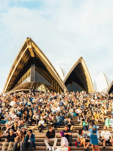 A large crowd sits on the steps outside the Sydney Opera House under a partly cloudy sky.