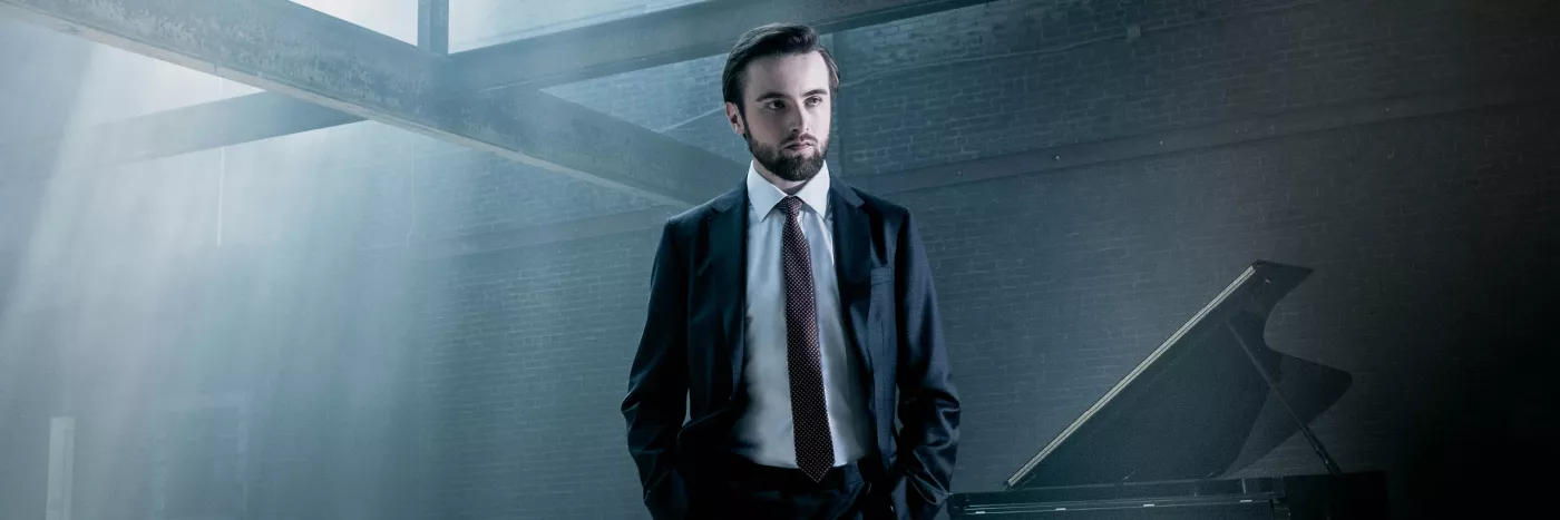 Pianist Daniil Trifonov stands in a suit in front of a grand piano