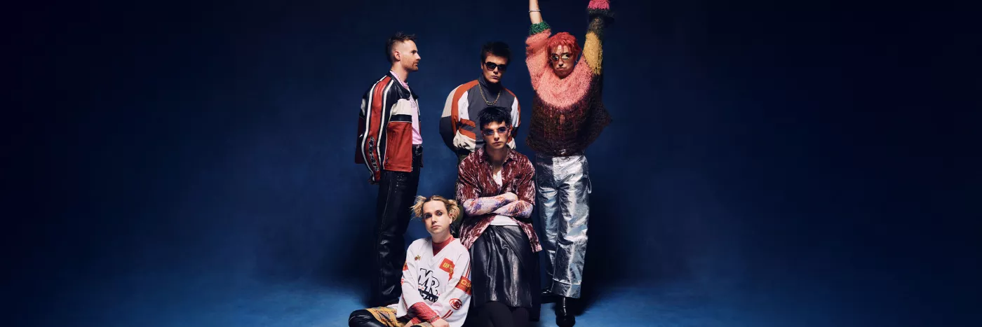 A group of five men dressed in kooky, punky Y2K outfits pose in front of a blue backdrop.